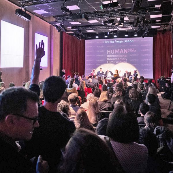 bilde av folkemengde som sitter med ryggen til og ser mot en scene med mennesker på paneldebatt.