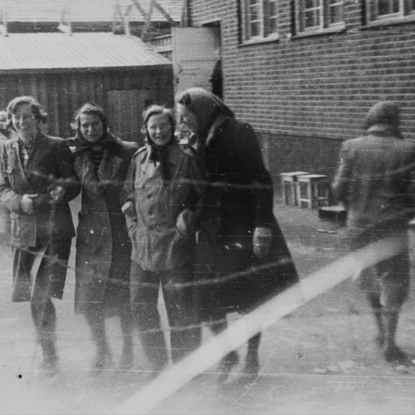 Fotografi av luftegård Grini fangeleir. Fire kvinner holder hverandre i armene og snakker sammen mens de går foran en murvegg. Foran i bilde ser vi piggtrådgjerde.